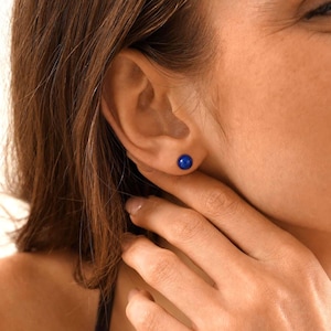 May include: A close-up of a blue stud earring in a person's ear. The earring is a small, round, and vibrant blue. The person's skin is fair, and their hair is brown. The earring is the focal point of the image.