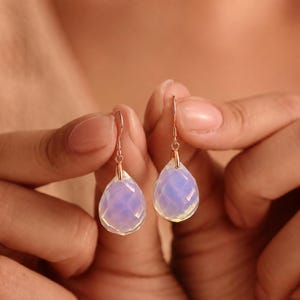 May include: A pair of drop earrings featuring faceted, pear-shaped opal stones with a translucent, iridescent blue and white glow. The stones are suspended from thin, rose gold-toned metal hooks that attach to the top of each gemstone.