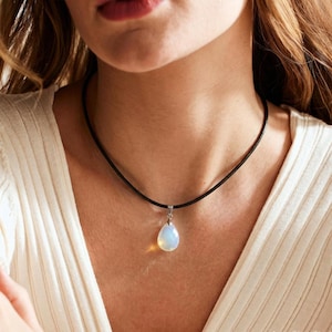 May include: A teardrop-shaped opal pendant hangs from a simple black cord necklace. The stone displays a milky, iridescent white sheen with subtle blue and yellow flashes. The pendant is secured by a small silver-toned metal bail at the top.