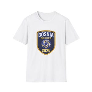 May include: A white short-sleeved t-shirt featuring a centered shield-shaped emblem. The emblem has a dark blue background with a gold border, displaying the text BOSNIA SOCCER 2026 and a stylized blue and yellow soccer ball graphic.