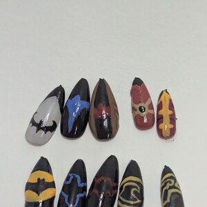 Peut inclure: Un ensemble de dix faux ongles en forme d'amande avec divers motifs peints à la main. La collection comprend des ongles noirs avec des symboles de chauve-souris jaunes, bleus et rouges, un ongle gris avec un logo noir, et des rouges avec des détails.