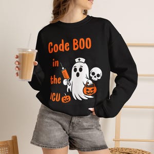 Könnte beinhalten: Ein schwarzes Langarm-Sweatshirt mit einer Grafik eines Geistes, der eine Krankenschwesterhaube trägt, eine orangefarbene Spritze und einen Kürbiseimer hält, sowie einen Totenkopf. Der Text lautet Code BOO in the ICU in orange.