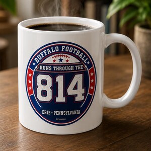 May include: A white ceramic mug filled with a dark hot beverage emitting steam. The front features a circular blue and red graphic with the text "BUFFALO FOOTBALL RUNS THROUGH THE 814 ERIE PENNSYLVANIA" and "2026 RJ" with stars and a football icon.