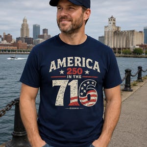 Op de afbeelding: Een marineblauw t-shirt met korte mouwen en een verweerde grafische print met de tekst AMERICA 250 IN THE 716, waarbij het getal 716 is vormgegeven met de strepen en sterren van de Amerikaanse vlag. Het ontwerp bevat de tekst ©2026 RJ onderaan.