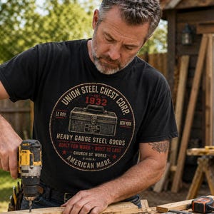 Könnte beinhalten: Ein schwarzes T-Shirt mit einem kreisförmigen Aufdruck mit dem Text "UNION STEEL CHEST CORP. 1932" und "HEAVY GAUGE STEEL GOODS". Ein Mann benutzt einen gelb-schwarzen Bohrer auf einem Holzbrett. Das T-Shirt ist in Amerika hergestellt.