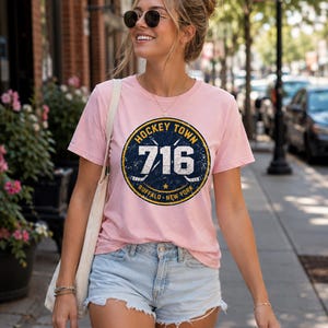 Op de afbeelding: Een lichtroze T-shirt met een marineblauwe en gele cirkelvormige afbeelding met de tekst "HOCKEY TOWN 716 BUFFALO NEW YORK". Het shirt wordt gecombineerd met een lichtblauwe spijkerbroekshort.