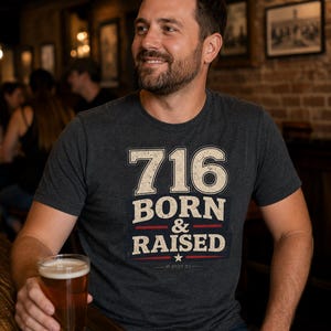 Op de afbeelding: Een antracietgrijs T-shirt met korte mouwen en de tekst 716 BORN & RAISED in een verweerd crèmekleurig lettertype, met een rode horizontale lijn en een klein sterretje. Het ontwerp bevat een donkerblauw rechthoekig element achter de tekst.