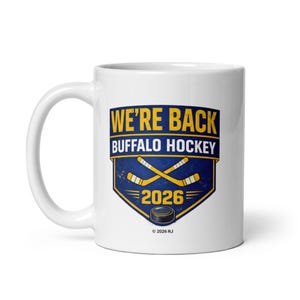 Puede incluir: Taza de cerámica blanca con un diseño temático de hockey. El diseño presenta el texto "WE'RE BACK BUFFALO HOCKEY" en amarillo y azul, junto con palos de hockey cruzados y un disco. También se muestra el año "2026".