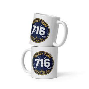 Op de afbeelding: Twee witte keramische mokken op elkaar gestapeld. Elke mok heeft een marineblauw en goud cirkelvormig ontwerp met de tekst "HOCKEY TOWN 716 BUFFALO - NEW YORK" en een hockeystick- en puck-afbeelding.