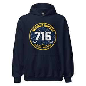 May include: A navy blue hooded sweatshirt featuring a circular graphic on the chest. The design includes the text BUFFALO HOCKEY, 716, and BUFFALO NEW YORK, along with two crossed hockey sticks and a small star, all in a distressed white and yellow print.