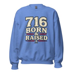 May include: A light blue crewneck sweatshirt featuring the text 716 BORN & RAISED in a distressed cream font with dark blue outlines. The design includes two horizontal red lines and a small white star, with the text 2026 RJ printed at the bottom.