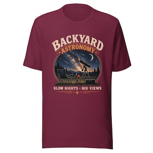May include: A maroon short-sleeved t-shirt featuring a graphic of a telescope, a folding chair, and a night sky with the Milky Way. The text reads BACKYARD ASTRONOMY at the top and SLOW NIGHTS • BIG VIEWS at the bottom in a cream-coloured font.