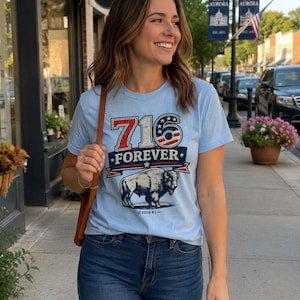 Op de afbeelding: Een lichtblauw T-shirt met korte mouwen en een grafische print met de tekst 716 FOREVER, een gestileerde illustratie van een bizon en rode, witte en blauwe sterren. Het ontwerp bevat onderaan de kleine tekst 2026 RJ.