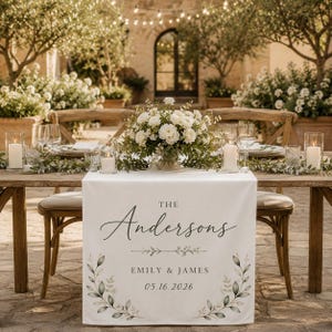 May include: A white fabric table runner featuring the text THE Andersons, EMILY & JAMES, and 05.16.2026 in black lettering. The design includes a botanical leaf illustration at the bottom and a decorative line element above the names.
