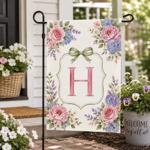 May include: A rectangular garden flag featuring a cream background with a large pink letter H centred inside a decorative frame topped with a sage green bow. The corners are adorned with watercolour-style pink roses and blue hydrangea clusters.