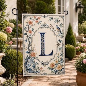 May include: A rectangular garden flag featuring a cream background with a large dark blue letter L centred in an ornate frame. The design includes a blue and white vase with pink flowers, green bamboo stalks, and a small blue bird perched on a branch.