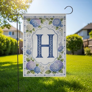 May include: A garden flag featuring a large blue letter "H" within a white and blue floral frame. The flag is adorned with blue and purple hydrangea flowers and green leaves, set against a light background. The flag is displayed on a black metal stand in a grassy yard.
