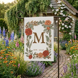 Peut inclure: Un drapeau de jardin rectangulaire présentant une lettre M centrale dans une police serif, avec le texte EST. 2025 en dessous. Le motif est encadré par une couronne botanique de roses rouges et orange, de feuilles vertes et de fines branches.