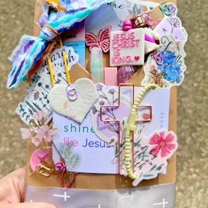 May include: A wrapped gift with a variety of decorative elements. The package is wrapped in brown paper and tied with a gray ribbon. Embellishments include a butterfly, cross, and the text "Jesus Christ is King."