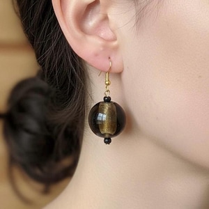 May include: A close-up of a gold-colored earring with a black sphere and gold accents. The earring is hanging from a gold-colored hook. The earring is round and has a gold stripe down the middle.