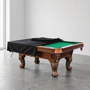 May include: A large, ornate wooden pool table with a green felt playing surface. A black cover is partially draped over the table. The table has carved legs and leather pockets. The room has a neutral colour palette.
