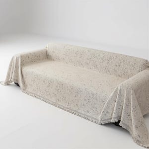May include: A beige sofa blanket with a speckled pattern covers a sofa. The blanket has a fringed edge and drapes over the arms and back of the sofa. The background is a plain white.