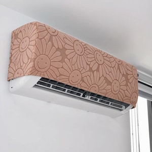May include: A rectangular air conditioner cover featuring a repeating pattern of dusty pink daisies with smiling faces. The fabric cover fits over the top and front of the white wall-mounted unit, leaving the horizontal air vents visible at the bottom.