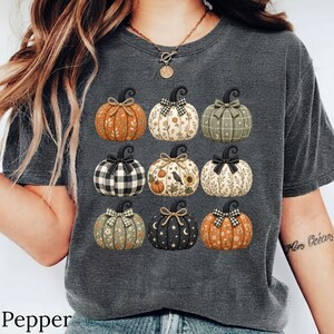 May include: A charcoal grey short-sleeve t-shirt featuring a graphic print of nine pumpkins arranged in a grid. Each pumpkin displays a unique pattern, including floral, plaid, and celestial designs, and is topped with a decorative fabric bow.