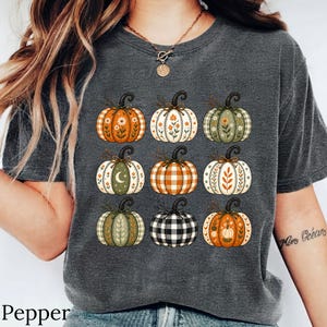 Op de afbeelding: Een donkergrijs t-shirt met een grafische print van negen pompoenen in een raster. De pompoenen hebben verschillende patronen, waaronder oranje en wit ruitpatroon, zwart-wit vichy, effen groen en bloem- of maanmotieven in aardetinten.