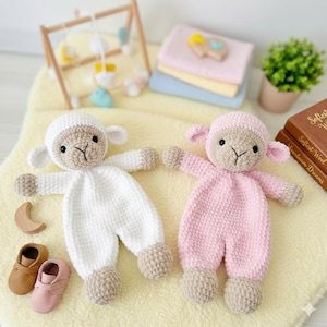 Könnte beinhalten: Zwei gehäkelte Lamm-Puppen, eine weiße und eine rosa, mit beigen Akzenten. Die Puppen liegen auf einer gelben Decke, mit Babyschuhen und einem Holzspielzeug. Ein Holz-Babyspielplatz und andere Babyartikel sind im Hintergrund.