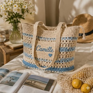 Peut inclure: Un sac fourre-tout en crochet beige et bleu avec le nom "Camille" brodé sur le devant. Le sac a des rayures bleues et un cœur bleu. Un chapeau de paille et un vase de marguerites sont en arrière-plan.