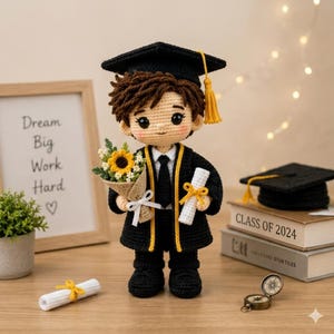 Peut inclure: Poupée de graduation crochetée, portant une toge et un chapeau noirs avec des détails jaunes, tenant un bouquet de fleurs et un diplôme. La poupée a les cheveux bruns et se tient devant un cadre avec l'inscription "Dream Big Work Hard".