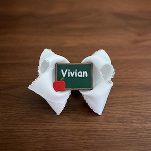 Peut inclure: Un nœud pour cheveux blanc orné d'un écusson rectangulaire vert foncé rappelant un tableau noir. L'écusson est brodé du prénom Vivian en lettres blanches et comporte une petite icône de pomme rouge dans le coin inférieur gauche de l'écusson.