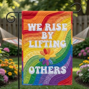 Może przedstawiać: Prostokątna flaga ogrodowa z pikowanym tęczowym tłem w faliste linie. Biały napis brzmi WE RISE BY LIFTING OTHERS, umieszczony nad kolorowym kwiatem z tkaniny o płatkach w różne wzory oraz odcieniach pomarańczu, żółci i błękitu.