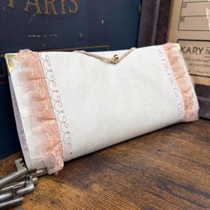 May include: A rectangular off-white paper envelope featuring delicate peach-colored ruffled lace trim on both sides. The envelope is secured with a thin string loop and a small gold-toned button closure, accented by decorative stitched patterns.
