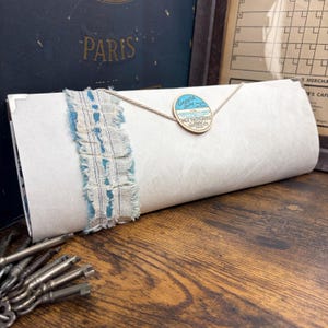 May include: A rectangular white paper envelope secured with a light-colored string and a circular blue and white Coopers Dairy label. A strip of frayed blue and white fabric is wrapped vertically around the left side of the envelope.