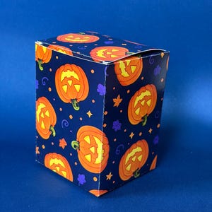 May include: A rectangular cardboard box featuring a dark blue background with a repeating pattern of orange jack-o'-lanterns, small purple leaves, and tiny yellow stars. The box has a folded top closure and displays a festive Halloween theme.