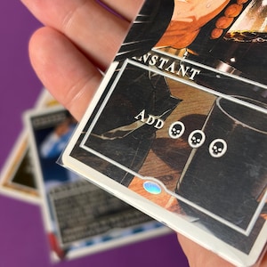 May include: A rectangular trading card in a clear protective sleeve featuring an illustration of a hand pouring dark liquid into a glass. The card text includes the word INSTANT and the phrase ADD followed by three black skull mana symbols.