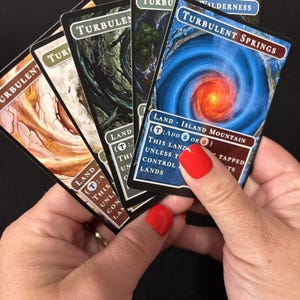 May include: A hand holding a fan of trading cards. The cards feature the word "Turbulent" and various landscape illustrations. One card reads "Turbulent Springs" with a blue and red spiral design. The cards are for a game.
