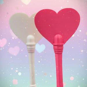 May include: Two heart-shaped hand fans, one white and one bright pink, are displayed against a soft, pastel background. The fans have long, decorative handles. The background features floating hearts and sparkling accents, creating a whimsical aesthetic.