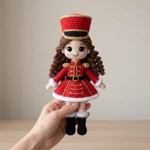 Puede incluir: Una muñeca de ganchillo vestida de soldado cascanueces, con chaqueta roja bordada en oro, sombrero rojo a juego con borde dorado, cinturón negro, borde de falda blanco y botas negras. La muñeca tiene el pelo largo, castaño y rizado.