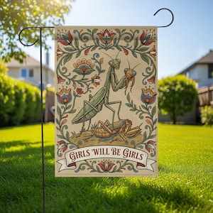 Puede incluir: Una bandera de jardín beige con una mantis religiosa verde sosteniendo un insecto más pequeño, con el texto "GIRLS WILL BE GIRLS" en una pancarta. La bandera tiene un borde floral con detalles en rojo, azul y verde. La bandera está en un soporte de metal negro.