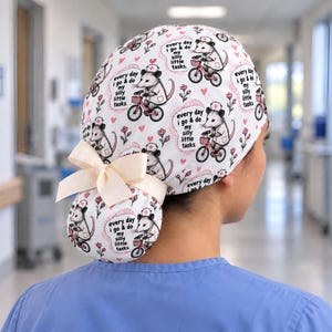 May include: A white scrub cap featuring a repeating pattern of opossums wearing nurse hats and riding bicycles, accompanied by the text "every day i go & do my silly little tasks." The cap includes small pink hearts, flowers, and a cream-colored ribbon bow.