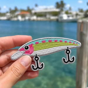 May include: An embroidered patch shaped like a fishing lure. The lure features a light green back with pink spots, a bright pink head with a black eye, and a white underbelly. Two black treble hooks are attached to the bottom of the lure body.