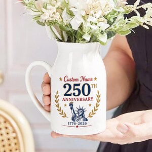 May include: White ceramic pitcher with a handle, decorated with the text "Custom Name 250th Anniversary 1776-2026" and a graphic of the Statue of Liberty. The pitcher is filled with white flowers.