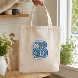 May include: A natural-colored canvas tote bag featuring a large, embroidered letter B in blue. The letter is decorated with white and blue floral embroidery, including daisies and smaller blossoms, with green stems and leaves along the left side.