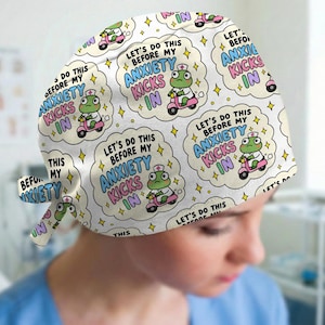 May include: A white scrub cap featuring a repeating pattern of a green frog wearing a nurse hat and riding a pink scooter. The design includes the text "LET'S DO THIS BEFORE MY ANXIETY KICKS IN" inside a cloud-shaped bubble, surrounded by yellow stars.
