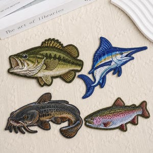 May include: Four embroidered fish patches featuring a green largemouth bass, a blue marlin, a dark brown catfish with long whiskers, and a multi-colored rainbow trout with pink and green scales, all displayed as detailed fabric appliques.