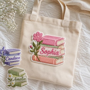 Peut inclure: Un sac fourre-tout en toile couleur crème avec un grand écusson brodé représentant des livres empilés en rose, vert et corail avec une fleur rose. Le prénom Sophia est brodé sur le livre central. Deux écussons supplémentaires avec Candace et Amelia.