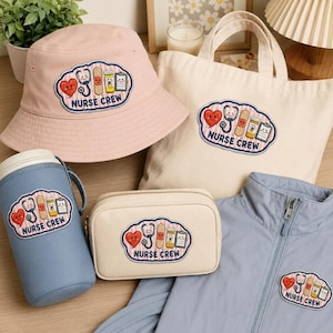 Può includere: Una collezione di articoli che include un cappello da pescatore rosa chiaro, una borsa in tela color crema, una custodia isolante blu per tazza, una pochette con cerniera color crema e una giacca azzurra, ognuno con una toppa con il testo NURSE CREW e icone mediche.